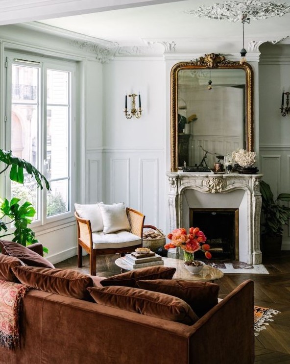 A sophisticated French chic living room with a French fireplace, a large mirror in an ornated frame, a neutral chair, a rust colored sofa with pillows and potted plants