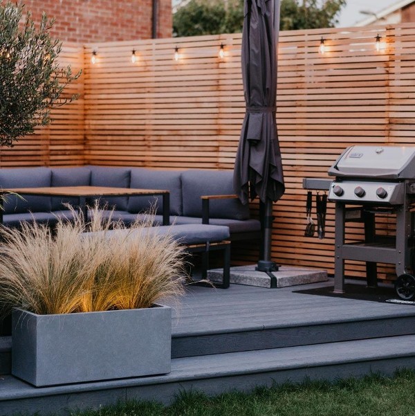 a stylish modern terrace with grain brushed basalt millboard decking, grey upholstered furniture, a wooden table, a concrete planter with lights and an umbrella