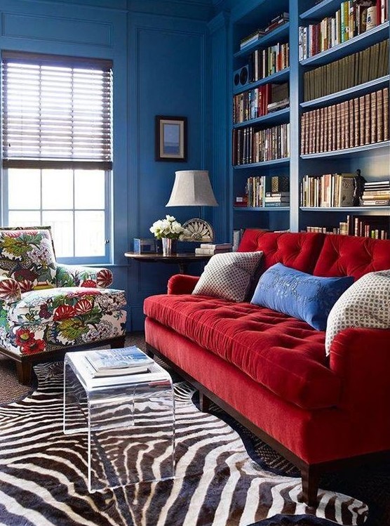 A vibrant space with blue walls, built in bookshelves, a red sofa and a floral chair, layered rugs and an acrylic table