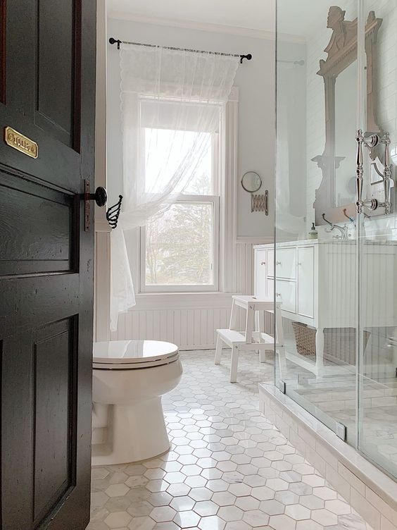 A white vintage bathroom with hex tiles and beadboard, a double hung window, a large vanity and a glass enclosed shower
