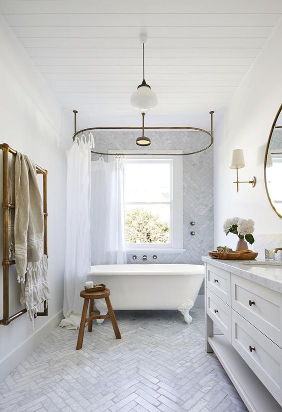 An airy bathroom with a double hung window, a large vanity, an oval clawfoot bathtub and a marble herringbone tile floor