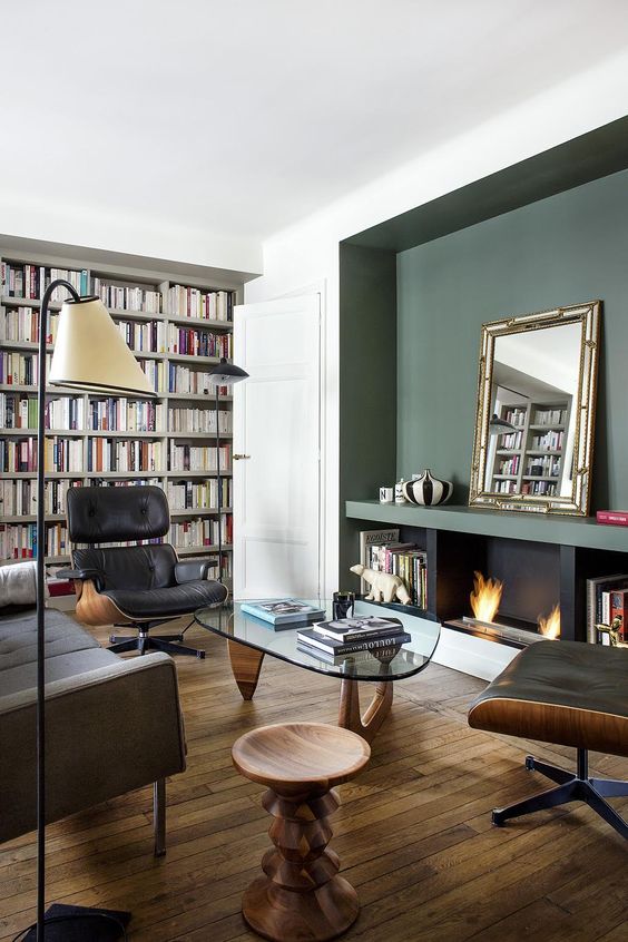 an elegant and sophisticated living room with a green accent wall, a fireplace, bookshelves taking a whole wall, a grey sofa, black Eames loungers and a glass coffee table