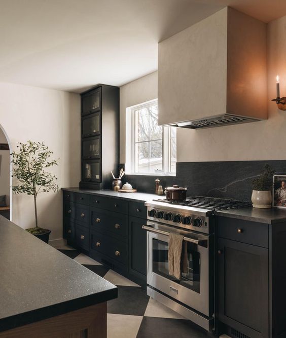 an elegant black and neutral kitchen with shaker style cabinets, black soapstone countertops and a backsplash, a hood and a checked floor
