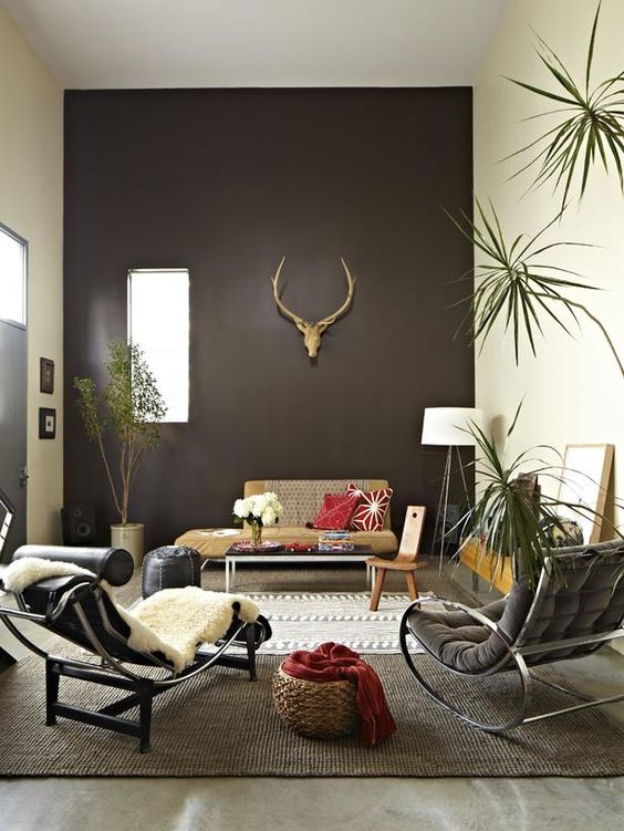 a catchy boho living room with a chocolate brown accent wall, a neutral sofa, a black and grey lounger, a chair and a pouf and some plants