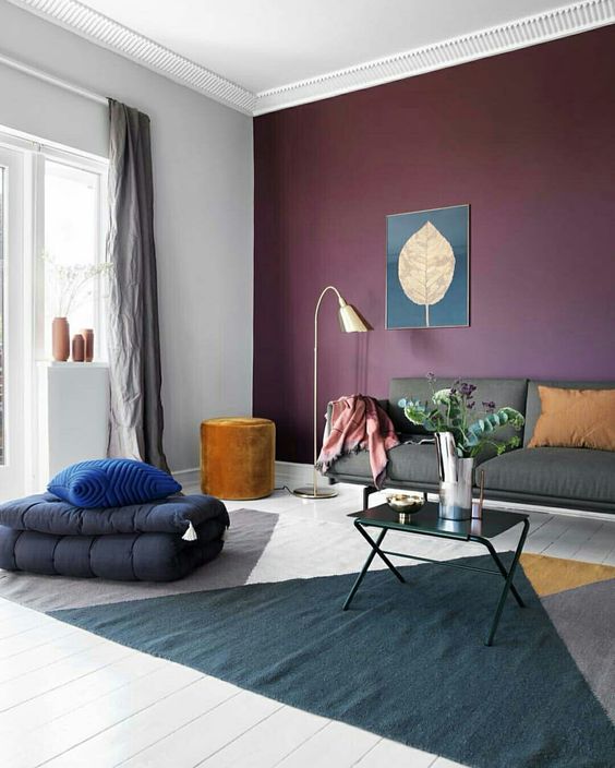 a catchy and bold living room with a purple accent wall, a grey sofa, a color block rug, black duvets, a gold lamp and a mustard pouf