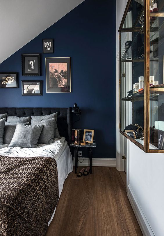 an attic bedroom with a navy accent wall and a gallery wall, a black upholstered bed, a glass shelving unit on the wall