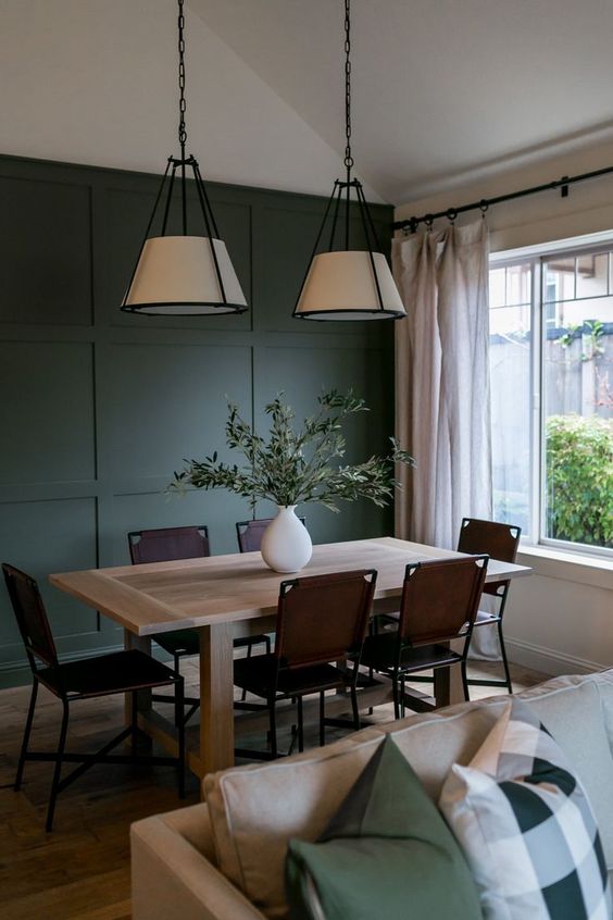 a cool dining room with a dark green paneled wall, a butcherblock table, brown chairs, pendant lamps and neutral curtains