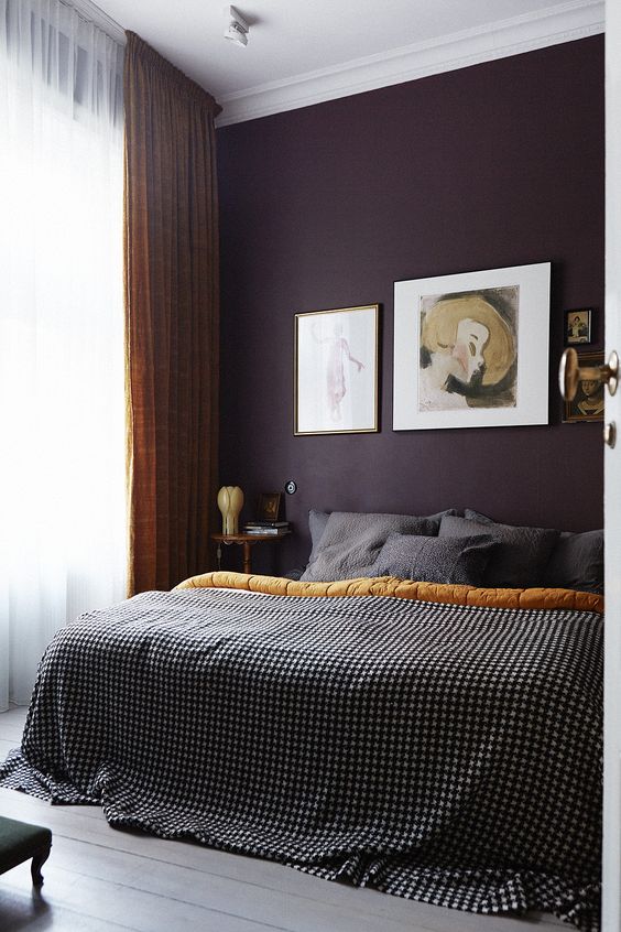 a refined and chic moody bedroom with a deep purple accent wall, a bed with grey bedding, nightstands, some artwork and mustard curtains