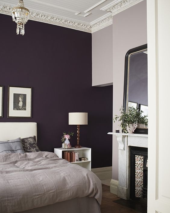 A chic bedroom with a deep purple accent wall, a white upholstered bed, grey bedding, a non working fireplace, a mirror and a crystal chandelier