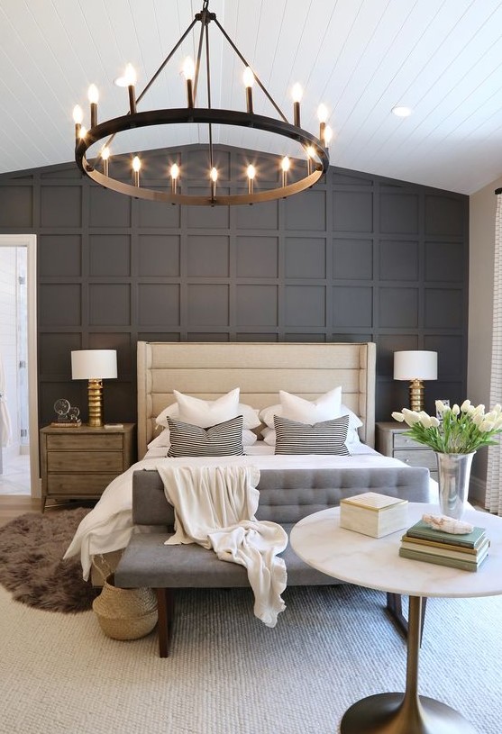 a stylish bedroom with a black paneled wall, an upholstered bed and bench, a round chandelier, wooden nightstands and layered rugs
