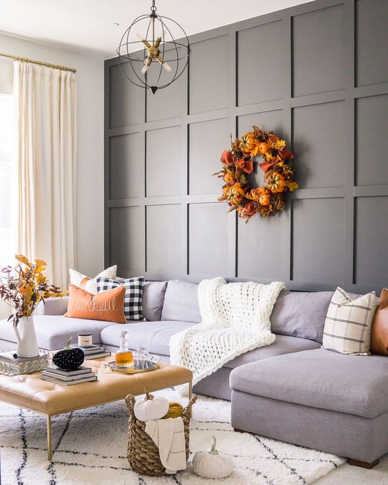 a cozy fall living room with a grey paneled wall, a grey sectional, a beige leather ottoman, some pretty season decor