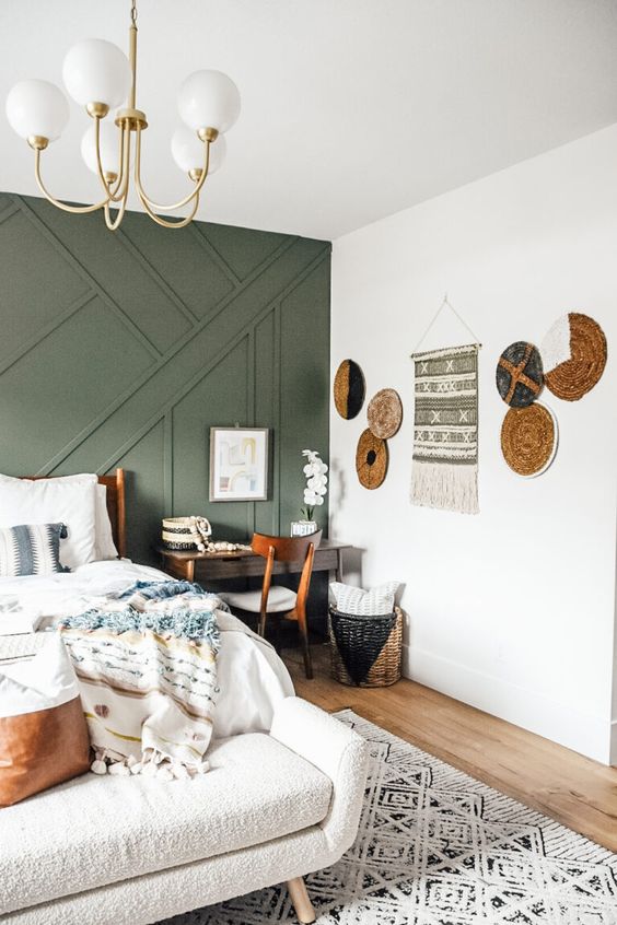 a cool boho bedroom with a green paneled wall, a bed with neutral bedding, a bench, a mini desk, a lovely gallery wall with platters