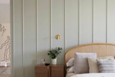 29 a peaceful bedroom with a pale green paneled wall, a rattan bed with neutral bedding, a stained nightstand and a gold sconce