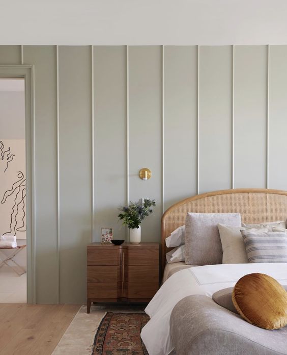 a peaceful bedroom with a pale green paneled wall, a rattan bed with neutral bedding, a stained nightstand and a gold sconce