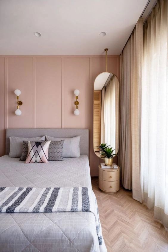a glam bedroom with a pink paneled wall, a grey bed with grey bedding, an oval mirror, gold sconces and neutral curtains
