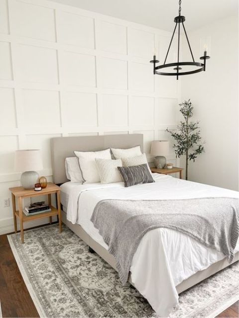 a neutral bedroom with a paneled accent wall, a grey bed with neutral bedding, stained nightstands, a printed rug and a chandelier
