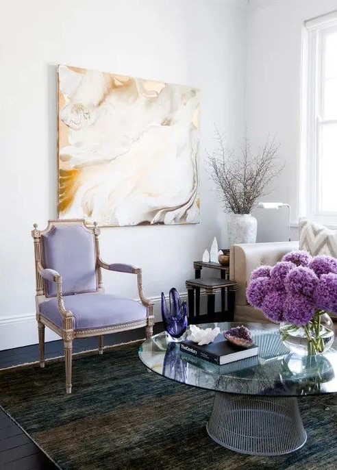 an airy refined living room with a lilac antique chair, a statement artwork, a dark rug and a glass coffee table with decor and flowers