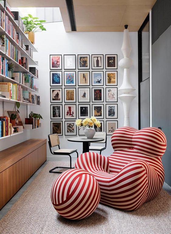 A lovely and bright nook with open bookshelves, a colorful gallery wall, a table and chairs, a bold catchily shaped red and white striped chair with a footrest
