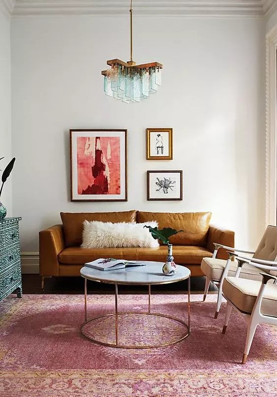 A colorful living room with a rust colored leather sofa, beige chairs, a round table, a pink rug and a blue inlay dresser, a whimsical mini gallery wall