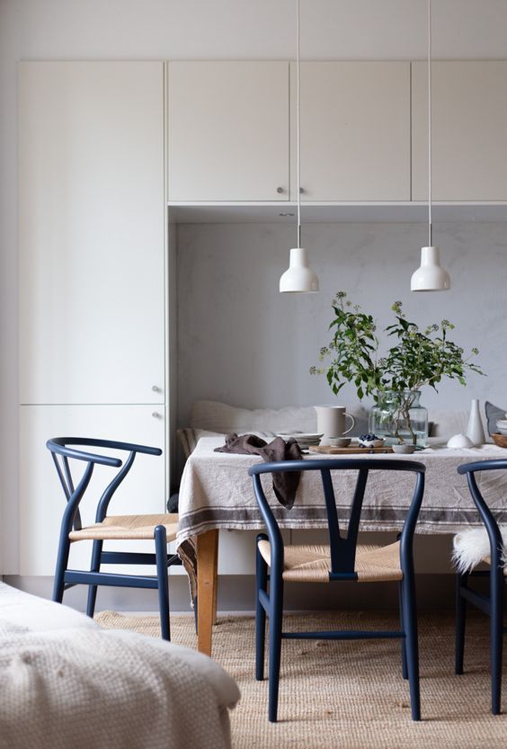 A Scandi dining space with a storage unit and a built in seat, a dining table and navy wishbone chairs, white pendant lamps