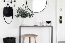 a Scandinavian black and white entryway with a black console table, a wooden stool, a round mirror, a rack, a basket and a rug
