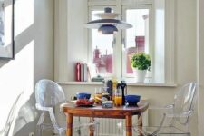 a Scandinavian breakfast nook with a stained table, ghost chairs, a Nordic pendant lamp and some books and potted greenery