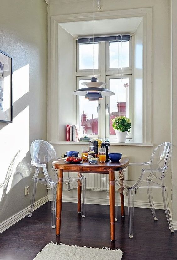 a Scandinavian breakfast nook with a stained table, ghost chairs, a Nordic pendant lamp and some books and potted greenery