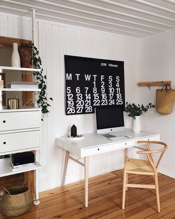 A Scandinavian dining space with a white desk, a light stained chair, a storage unit, a calendar and some greenery