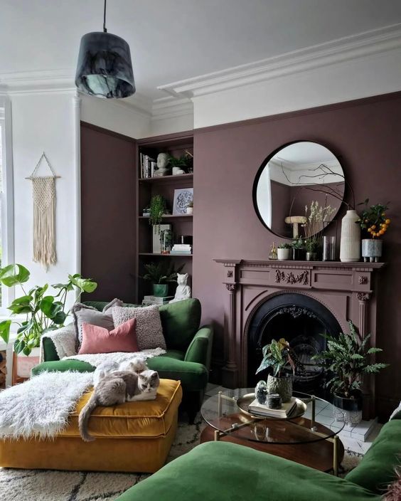 a beautiful boho living room with a purple wall, a non-working fireplace, built-in shelves, a green chair and a sofa, a mustard ottoman and lots of potted plants