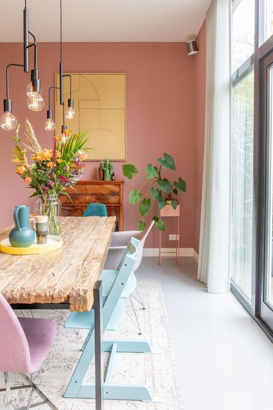 a beautiful dining room with a dusty pink accent wall, a rough wood dining table, pastel chairs, black bulb pendant lamps