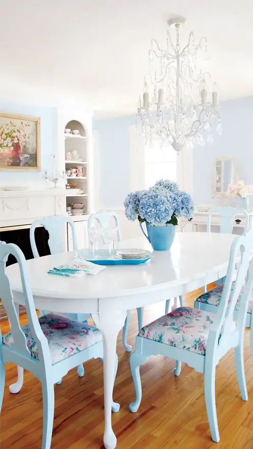 a beautiful pastel blue and white dining room with a pale blue accent wall, a fireplace, an oval table, blue chairs with floral upholstery and a crystal chandelier