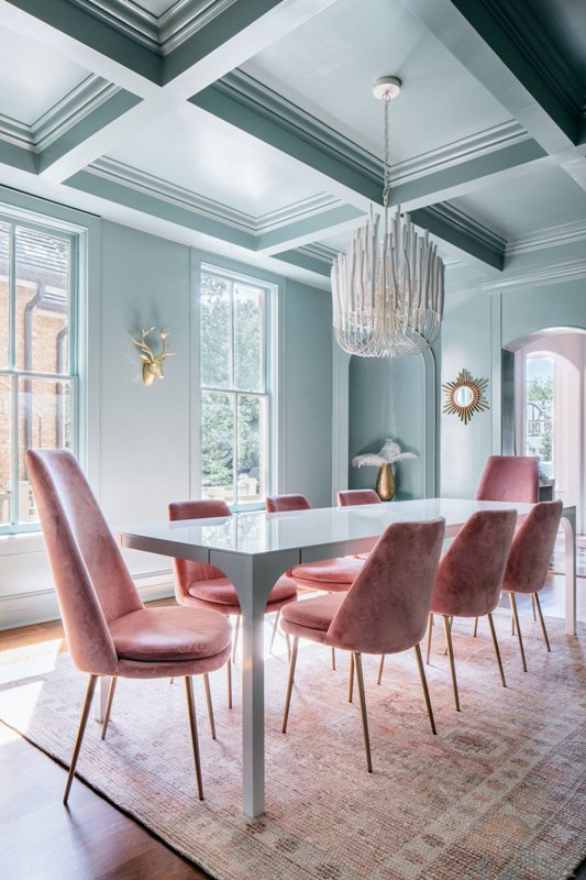 a beautiful pastel blue dining room with a coffeered ceiling, large windows, a corner niche, a blue table and pink chairs