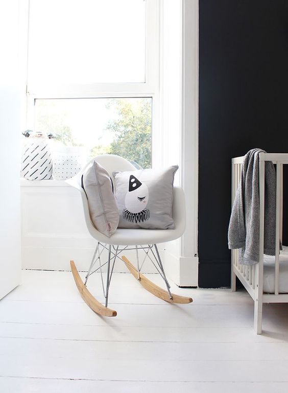 a black and white Scandinavian space with a black accent wall, a white crib with white bedding, a white Eames rocker and printed pillows