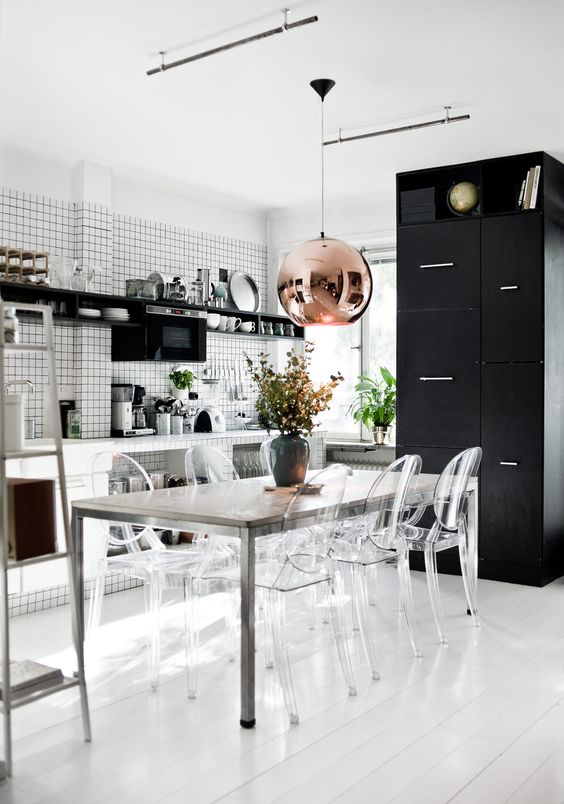 a black and white eat-in kitchen with white open cabinets, white tiles with black grout, a metal dining table and ghost chairs plus a copper pendant lamp