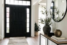 a black and white entryway with a black door, a printed rug, a console table, a round mirror and greenery