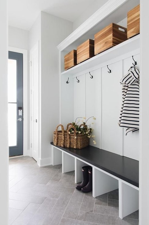 A black and white farmhouse mudroom with a built in shelf, a bench, baskets and boxes and a black door