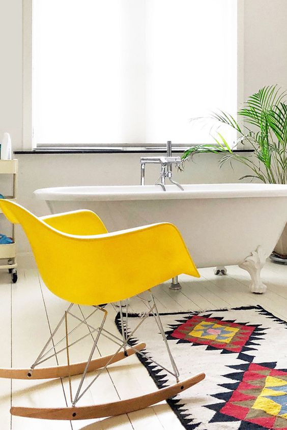 a boho bathroom with a free-standing bathtub, a yellow Eames rocker, a bold boho rug and some greneery
