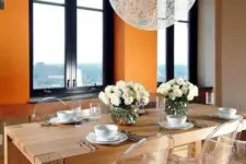 a bold dining room with orange walls, a stained table, ghost chairs, a woven pendant lamp and cool views