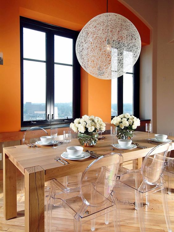 a bold dining room with orange walls, a stained table, ghost chairs, a woven pendant lamp and cool views