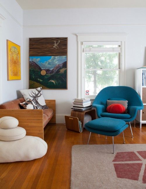 A bold mid century modern living room with rust colored loveseat, a blue Wumb chair with an ottoman, a curved shelf and pillows