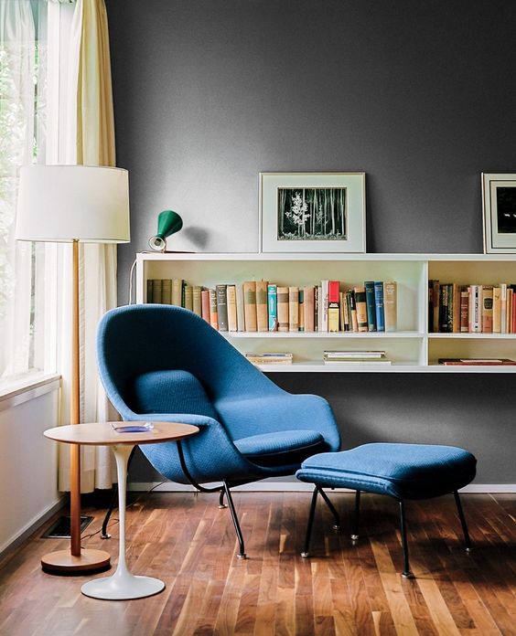 A bold mid century modern reading space with a blue Wumb chair and ottoman, a side table and a bookshelf on the wall