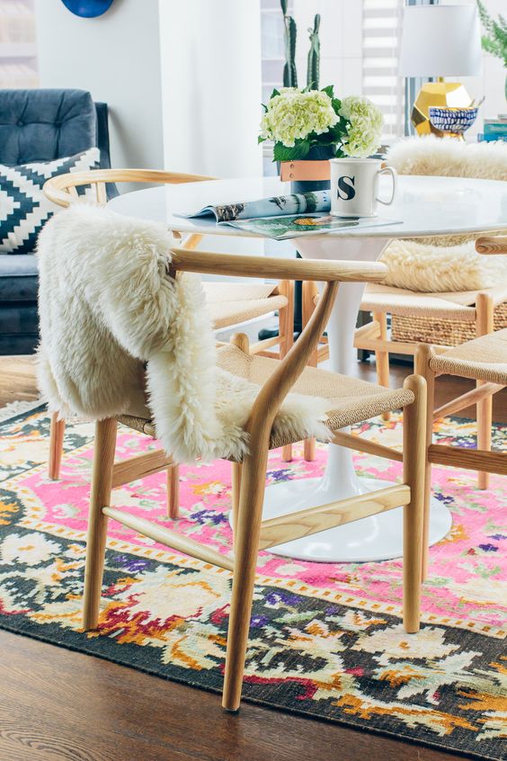 A bright dining room with a round table, light stained wishbone chairs, a colorful rug and soem faux fur