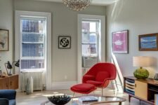 a bright living room with grey walls, a blue sofa and chair, a hot red Wumb chair and ottoman, a coffee table and a stained credenza