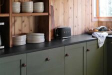 a cabin kitchen with stained wood walls, olive green lower cabinets, black stone countertops and a sconce