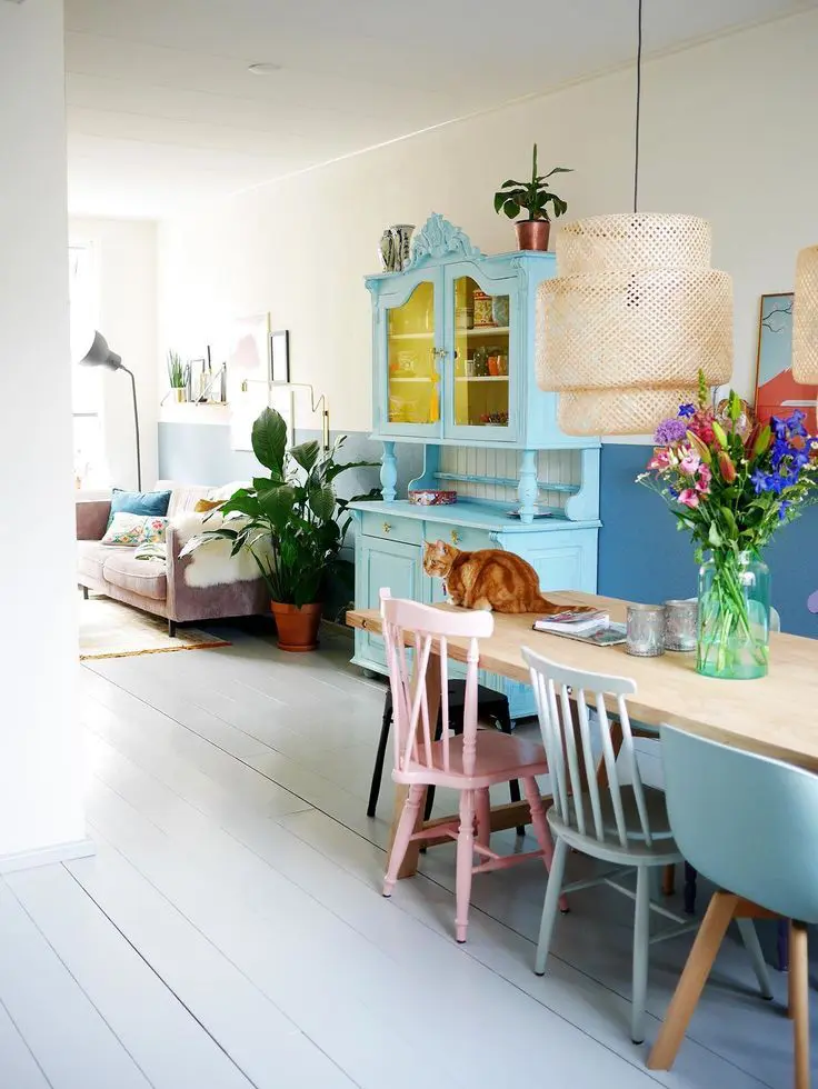 a catchy dining space with a long dining table, mismatching pastel chairs, a blue accent wall, woven pendant lamps and a blue vintage buffet