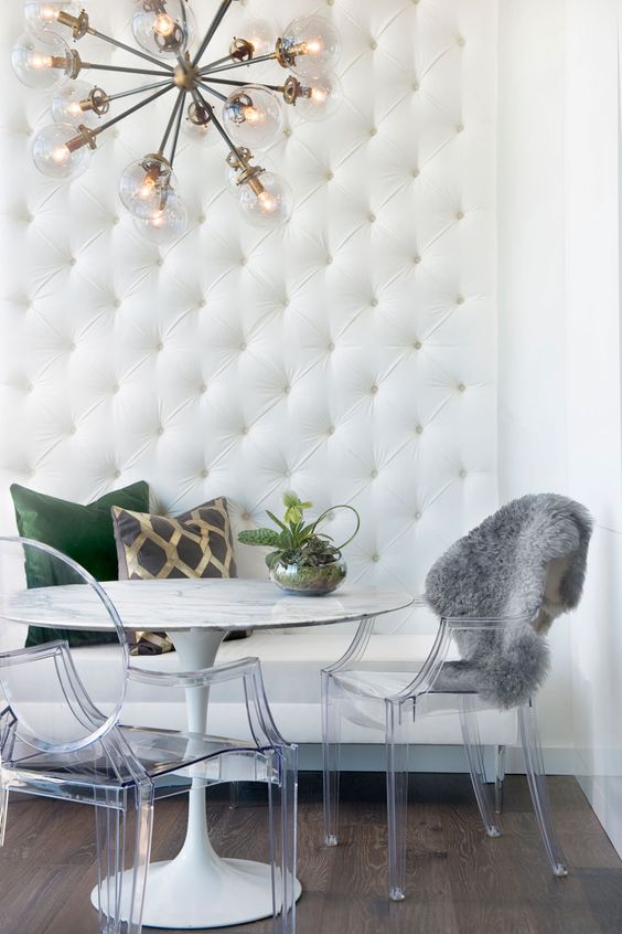 a catchy glam dining space with a built-in white leather bench, a round table, ghost chairs, a burst chandelier and some pillows