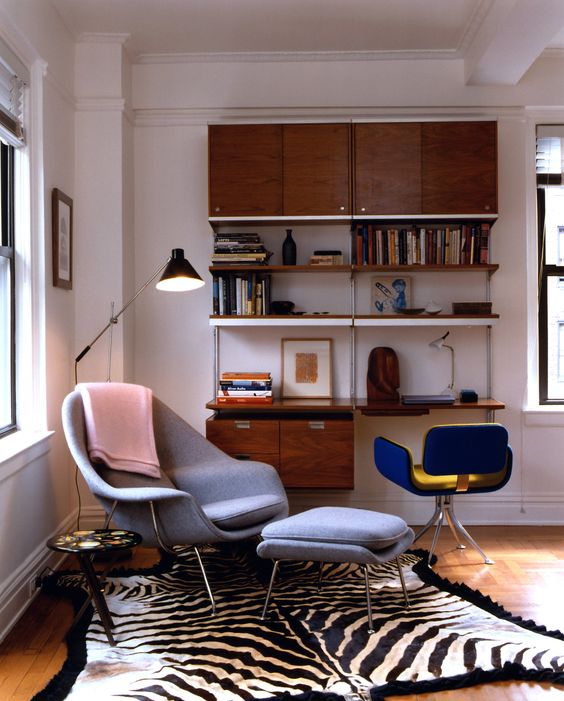A catchy mid century modern living room with a stained wall mounted shelving unit, a blue chair, a grey Wumb chair and ottoman, a printed rug
