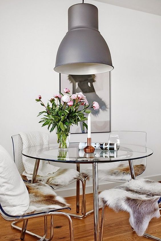 a catchy modern dining space with a round glass table, ghost chairs with faux fur and pillows, a grey pendant lamp