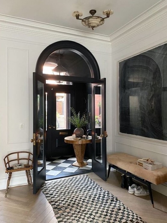 a chic black and white entryway with a neutral bench, a stained chair, a black and white rug and black doors