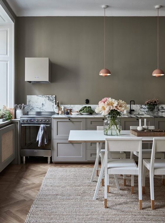 a chic olive green kitchen with grey cabinets, a small hood, pink pendant lamps, a white dining table and chairs